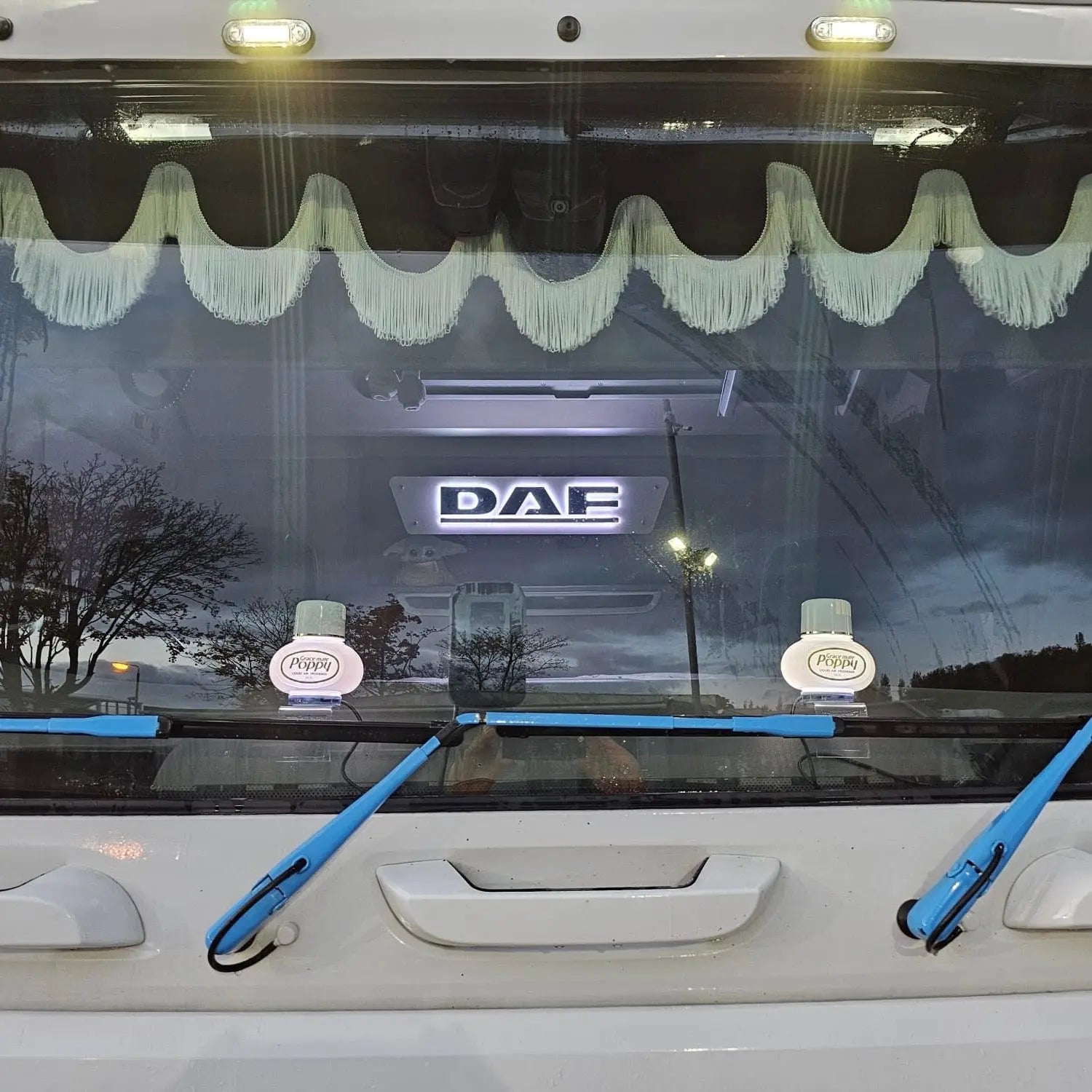 DAF 3D Truck Light Board on truck windshield, featuring an illuminated badge, cream fringe valance, Poppy air-fresheners, and blue wiper arms.