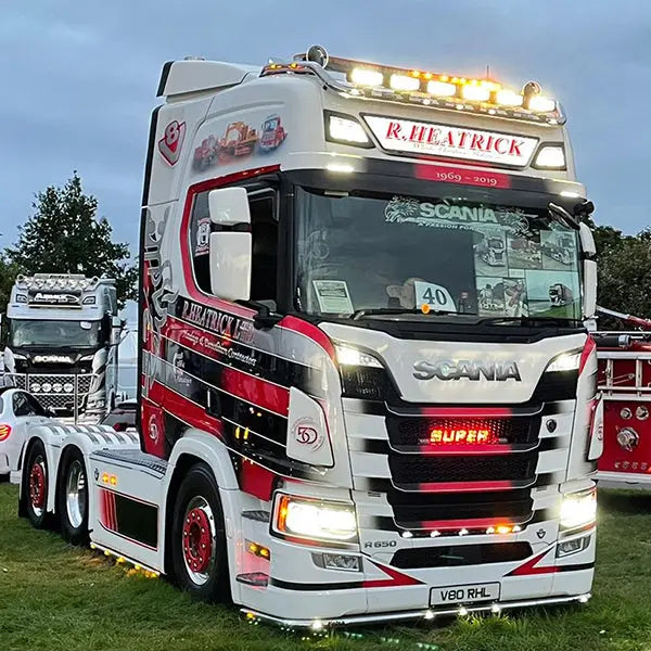Scania R650 V8 with 'SUPER' 3D Light badge, visible on the illuminated grille, at a truck show, showcasing customized chrome elements and R. HEATRICK livery.