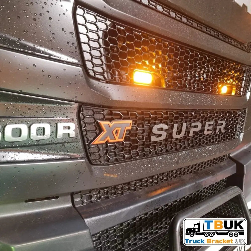 Close-up of a black honeycomb grille featuring a Next Gen R/S Series Grill Beacon/Strobe Light Mounting Bracket, fits Scania, with illuminated amber marker lights and water droplets.